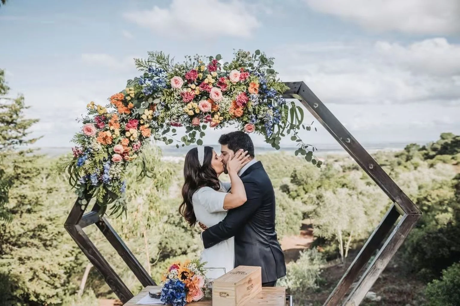 Noivos beijam-se sob um arco de flores com paisagem ao fundo